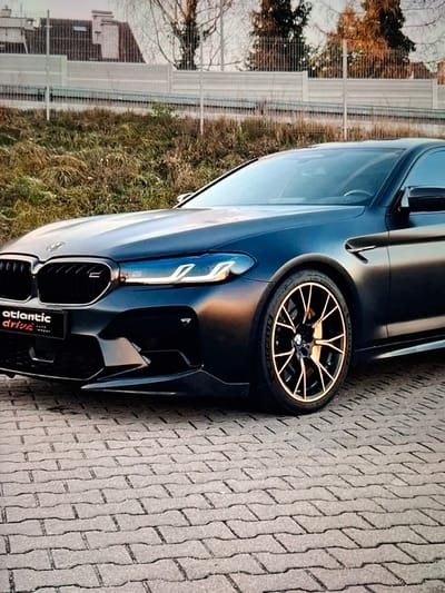 The image shows a close-up, low-angle view of a black BMW M5 sedan parked on a brick-paved surface. The car's front grille, headlights, and a portion of the hood are visible. The headlights have a distinctive daytime running light signature. The car has aftermarket wheels with a copper or bronze finish. A small black badge with white text "ATLANTIC DRIVE" is affixed to the lower left of the grille. In the background, a dry, grassy embankment is visible behind a metal fence. Beyond the embankment, there are bare trees and a building with a sloped roof. The lighting suggests it is either early morning or late afternoon. There are no people or actions depicted in the image. The scene is an outdoor setting, likely a parking area or roadside. The location is not Urus-Martan, Russia.