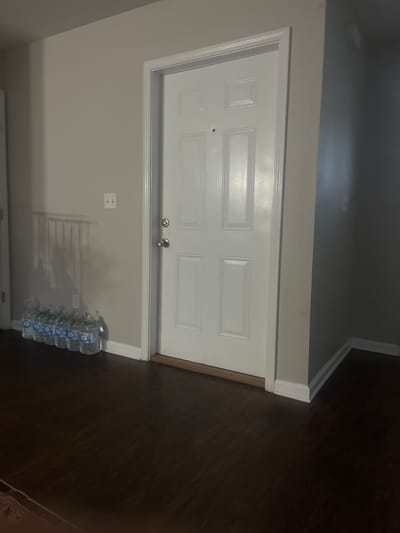 A white paneled interior door is positioned in the center of the frame. To the left of the door, a row of clear plastic water bottles with blue labels are arranged on the dark wooden floor. A light-colored wall with a visible light switch is to the left of the door. The floor is a dark brown wood laminate. The scene appears to be indoors, likely in a residential setting.