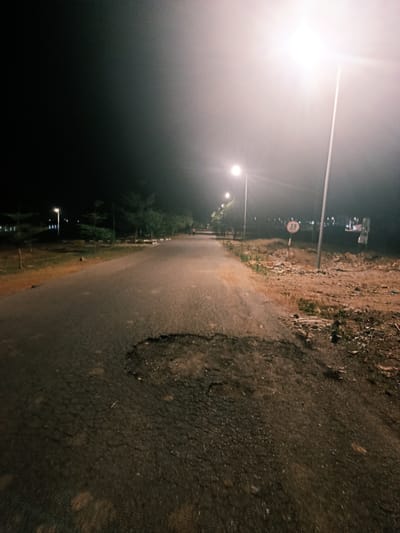 The image depicts a dimly lit road in Rafin Pa, Nigeria at night. Streetlights illuminate the road, which is paved but has a large pothole in the foreground. The roadside consists of vegetation and bare dirt. A speed limit sign displaying "25" is visible next to a streetlight on the right side of the road. Trees line the left side of the road. A person is walking in the distance.