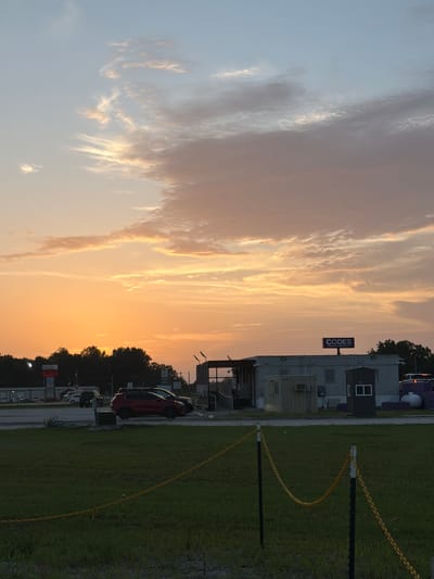 This image captures a sunset scene in Cardwell, United States. The sky is dominated by hues of orange, yellow, and pink, with scattered clouds. In the foreground, a grassy field is sectioned off by a yellow chain attached to black posts. Beyond the field, a parking lot is visible with several vehicles, including a red SUV and a black SUV. A long, grey mobile building complex stands in the middle ground, with a small guardhouse or booth to its right. A prominent sign with the word "CODES" in large purple letters and "CANNABIS DISPENSARY" in smaller white letters is mounted on top of the building. In the background, a line of trees separates the foreground from the sky. The overall impression is a commercial area at dusk.
