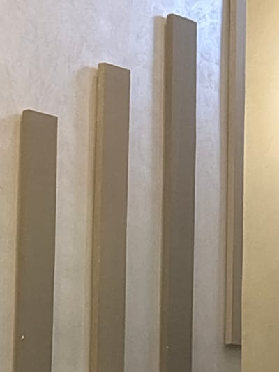 The image shows a close-up of a textured wall with three vertical, beige-colored decorative moldings. These moldings are of varying heights and widths, with the shortest and widest on the left, followed by a taller and narrower one, and then the tallest and widest on the right. To the right of the moldings, a portion of another beige decorative trim is visible. The wall itself appears to have a subtle, uneven texture. The lighting suggests an indoor environment, with shadows cast by the moldings indicating a light source from the right. No people or specific landmarks are identifiable, and the context of Berrechid, Morocco, cannot be inferred from the visual information alone.