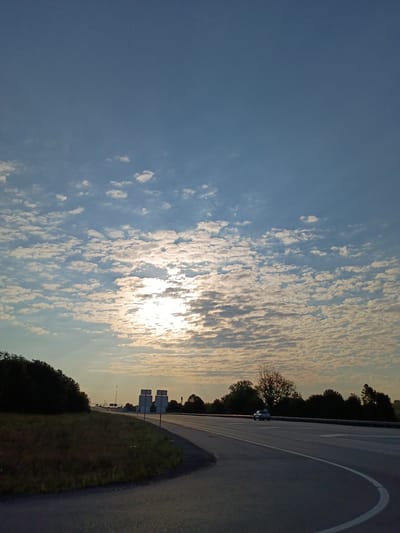 The image depicts a wide-angle view of a highway scene during sunrise or sunset. The sky is filled with numerous small, wispy clouds, creating a textured pattern across the blue expanse. The sun is partially obscured by these clouds, casting a warm, soft glow that illuminates the scene.

In the foreground, a paved highway curves to the right. The road surface is asphalt, marked with white lines. To the left of the highway, a grassy embankment slopes upwards and is covered with dark green foliage. Further along the highway, on the right side of the road, a single white car is visible driving away from the viewer. The background features a line of trees and vegetation, silhouetted against the sky. In the distance, several road signs with white surfaces are present.

The image does not contain any specific landmarks or visual cues that would definitively place it within the city of Richmond, United States of America.