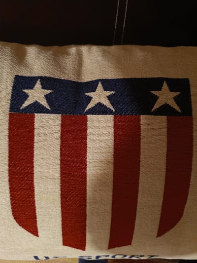 This is a close-up, slightly angled shot of a pillow or cushion with a design inspired by the American flag. The design features a blue band across the top with three white stars. Below the blue band are alternating vertical stripes of red and off-white. The texture of the fabric is visible, appearing somewhat rough and woven. The lighting appears to be artificial, casting soft shadows and highlighting the texture of the material. The background is dark and out of focus, suggesting the pillow is on a piece of furniture such as a couch or bed. At the very bottom edge of the image, partial text is visible in blue, appearing to be "U S PORT".