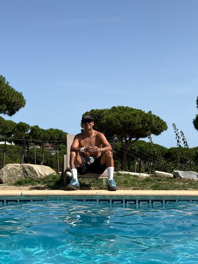 A shirtless man wearing black shorts, white socks, and blue sneakers sits on a lounge chair by a swimming pool. He is wearing a black baseball cap and sunglasses, and has a gold chain around his neck. He holds a clear glass with a beverage in his right hand. The man is outdoors with lush green trees and a clear blue sky in the background. Large rocks and grass surround the pool area. The scene appears to be a private residence in Sant Vicenç de Montalt, Spain, likely a backyard with a pool.