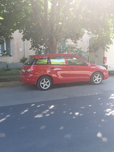 A red Toyota Matrix is parked on the side of a street in Dubossary, Moldova. The car is facing right in the frame. The vehicle is a station wagon with tinted windows. The car has five-spoke alloy wheels. There is a white van parked behind the red car. The street is paved with asphalt, and there is a sidewalk to the left of the car. The sidewalk is lined with grass and a small tree. In the background, there is a building with a light-colored facade and a window. The scene is bathed in bright sunlight, with dappled shadows from trees falling on the street and the car. The license plate on the red car is partially visible and reads "ZUH 454". The license plate on the white van reads "SZS 033".