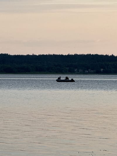 The image captures a serene lake scene in Glebychevo, Russia, during the soft light of dusk. A small, dark inflatable boat is positioned in the middle distance on the calm water, carrying three individuals. The boat appears to be moving, indicated by subtle ripples in its wake. In the background, a dense, dark green forest forms a treeline that stretches across the horizon, punctuated by a few distant buildings nestled amongst the trees. The sky above is a pale, muted gradient of pink and cream, suggesting the last moments of daylight. The overall atmosphere is tranquil and quiet, evoking a sense of peaceful solitude.