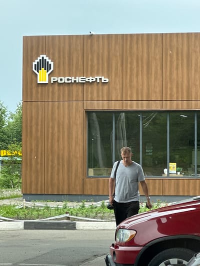 A man walks in front of a building with a "Rosneft" logo. The building has a wooden exterior and large windows. The man is wearing a grey t-shirt and black pants, and he is carrying a black bag over his shoulder. A red car is parked in front of the building. The scene appears to be a gas station or a service station. The image is taken in Slyudyanka, Russia, as indicated by the signage. The sky is overcast.