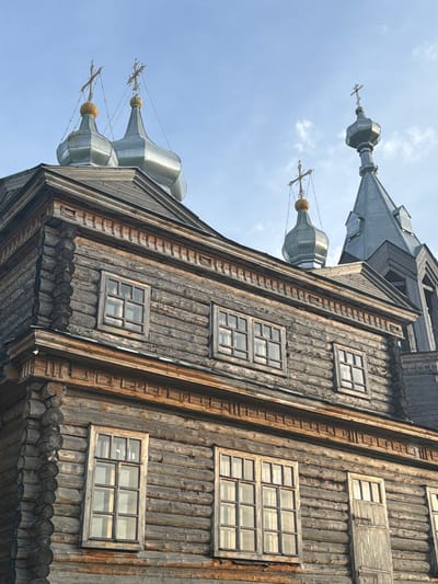 The image shows a detailed view of a historic wooden church with multiple onion domes topped with crosses, set against a bright blue sky with scattered clouds. The church is constructed from horizontal logs, weathered to a dark grey and brown hue. Intricate wooden carvings adorn the eaves and window frames, adding decorative detail to the rustic architecture. Several multi-paned windows are visible, reflecting the light. One door, also made of weathered wood, is present on the lower level. The perspective is from a low angle, looking up towards the domes and the upper sections of the church. The scene is devoid of people. This appears to be the Church of the Resurrection of Christ in Cherdyn, Russia, known for its traditional wooden architecture.