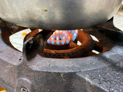 The scene, located in Sidi Chahmi, Algeria, presents a close-up view of a cooking setup. A large, circular, reflective metallic pot, likely aluminum or stainless steel, is positioned over a dark, heavy-duty cast-iron gas burner. Vivid blue flames are visibly emanating from the burner's orifices, directly heating the underside of the pot. The cast-iron burner exhibits signs of use, including some residue and surface texture. Partially visible behind the burner and beneath the pot, a fried egg with a distinct white albumen and yellow yolk rests on a lighter surface. No individuals or direct interactions are visible within the frame.