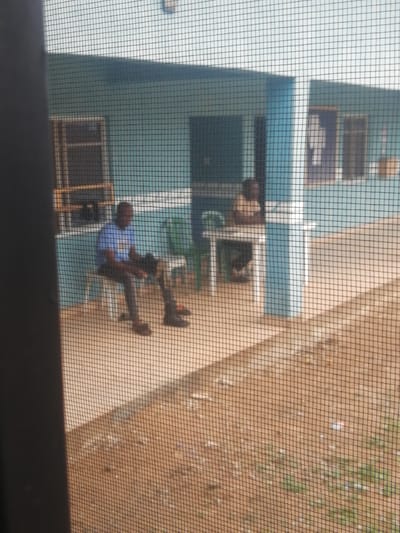 The image shows a view through a window screen of two men outside a light blue building, likely in Ajasse Ipo, Nigeria. One man, wearing a blue t-shirt and dark pants, sits on a white chair on a concrete patio, holding a dark object in his hands. Another man, wearing a light-colored shirt and dark pants, sits at a white table with a green chair next to it. The building has windows with dark frames and the ground outside is a mixture of dirt and scattered debris. The lighting suggests it is daytime.