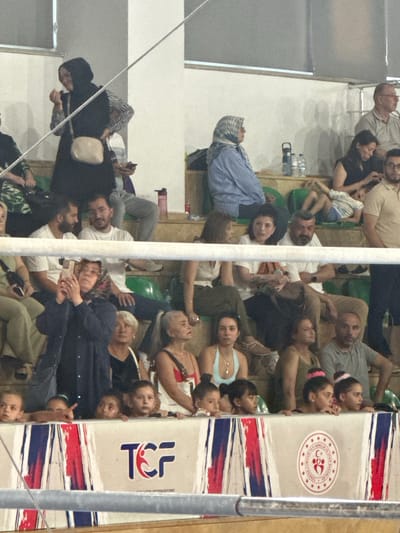 The image shows a crowd of people sitting in bleachers at an indoor sports venue, likely in Sancaktepe, Turkey. The focus is on the audience, with a few individuals in the foreground and more in the background.

In the foreground, a woman wearing a dark jacket and a headscarf is holding up a smartphone, appearing to take a picture or video. To her left, several children are visible behind a barrier, looking towards an unseen event. Their faces are a mixture of curiosity and focus.

The bleachers themselves are visible, with some seats colored green and others appearing to be wooden. There are barriers separating different seating sections. A large banner with "TCF" and what appears to be a logo is prominent in the lower part of the image.

In the background, a variety of individuals are seated, including men, women, and some children. Some are casually dressed, while others wear more formal attire. A woman in a blue striped shirt and headscarf is seated in the middle ground, looking to the left. Another woman with a dark headscarf is visible to the left of her. Further back, a man in a light-colored polo shirt is looking towards the right.

The overall atmosphere appears to be one of a sporting event or competition, with the audience engaged and attentive. The lighting suggests an indoor setting with artificial lighting. The presence of the "TCF" banner and the Turkish emblem on the lower banner suggests a national or Turkish-affiliated sporting event.