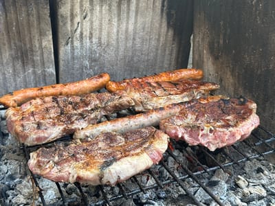 The image depicts a barbecue grill with several sausages and cuts of meat cooking over hot coals. The meat and sausages are arranged on a black metal grill rack. The background consists of textured, possibly concrete walls. The location is not discernible from the image alone.