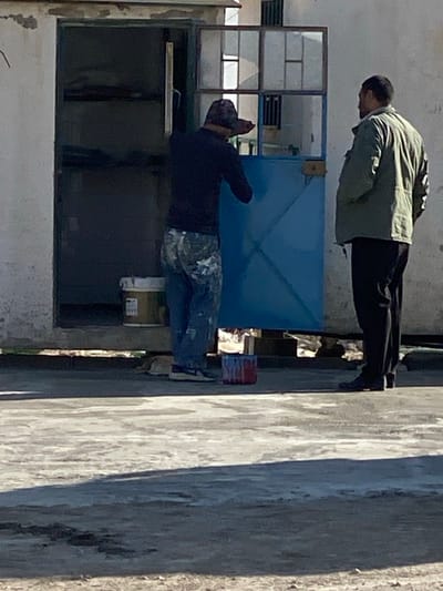 Two men are visible, one standing above the other. The upper man is wearing a light green jacket and black pants. The lower man is clad in a dark shirt and camouflage pants, his hands raised near his head. They are positioned near a window and blue metal structures, possibly associated with a building under repair. A concrete wall is seen to the right of the scene.