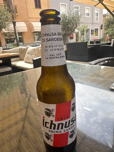 A close-up, eye-level shot focuses on a frosted, amber glass beer bottle of Ichnusa beer, a Sardinian brand, sitting on a reflective glass-topped table. The bottle's label is visible, featuring red and white elements with text and illustrations. Behind the bottle, the scene unfolds in an outdoor seating area of a restaurant or cafe, likely in Crabonaxa/Villasimius, Italy, given the beer brand. In the background, a building with awnings and windows is visible, along with trees and outdoor furniture such as chairs and sofas. Sunlight illuminates the scene, casting shadows and creating a bright, daytime atmosphere. The overall impression is that of a relaxed outdoor dining or drinking experience.