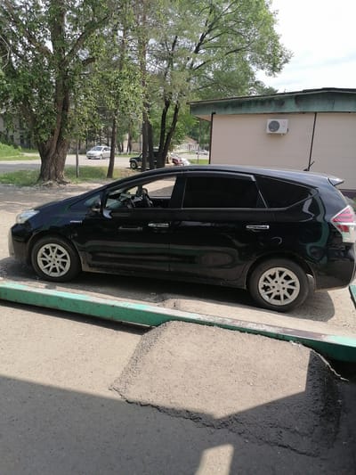 In Novopokrovka, Russia, a black Toyota Prius hybrid is parked on a paved surface adjacent to a building with an LG air conditioning unit mounted on its exterior. The car is positioned with its side facing the viewer, and its wheels are visible. In the background, there are trees, other vehicles, and a building. The scene is outdoors and appears to be during the daytime.