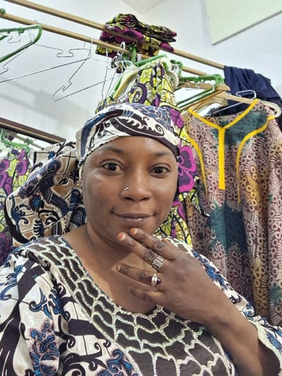 An adult female is depicted indoors in Gyadi Gyadi Arewa, Nigeria, looking directly at the camera. She possesses a medium skin tone, brown eyes, and a small metallic stud piercing on her right nostril. Her head is wrapped in a fabric headcover exhibiting a white, black, and brown geometric pattern with blue and gold accents. She is attired in a garment featuring a dark brown, white, and blue patterned fabric with an elaborate, light-colored, possibly silver-toned, embellished neckline. Her right hand is positioned beneath her chin; a silver-colored ring with an intricate design is visible on her index finger, and a ring with a dark red or brown stone is on her ring finger. A reddish-brown stain, consistent with henna, is present on her right fingertips and nails. The background reveals a light-colored wall and a horizontal clothing rail. Numerous garments, primarily composed of vibrant patterned fabrics (Ankara), hang from this rail using green plastic and silver wire hangers. Additional folded fabrics are stacked on top of the rail, and a section of dark blue fabric is visible on the far right. A partial wooden frame is visible on the left side of the background.
