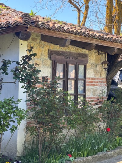 The image depicts a building in Kalimantsi, Bulgaria. The building has a tiled roof with wooden beams. The walls are constructed of stone and brick. There's a wooden-framed window with multiple panes. A bush with green leaves and red flowers is in front of the building. There is a tree branch above the building. The wall is painted white.