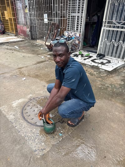 A man kneels on a wet concrete ground in what appears to be a street in Lagos, Nigeria. He is wearing a dark blue short-sleeved shirt with a subtle pattern and blue jeans. His hands are outstretched, with his right hand holding a green watering can. The watering can is positioned over a metal manhole cover, and it appears he is either filling it with water from the ground or emptying it onto the manhole.
The ground is wet and has puddles, suggesting it might have recently rained or there's an ongoing water source. Several black and green flip-flops are visible on a patterned tiled surface to the left of the man, near a building entrance. The building behind him has white bars or grates on its windows and doors, characteristic of many buildings in urban Nigeria. Some items are visible through the grates, including what looks like clothing and possibly fans. There is some debris and graffiti on the building walls and the ground.
The man's expression is one of focus. The time of day is unclear, but the lighting suggests it is daytime. The overall scene depicts a moment of daily life, possibly related to water management or a household chore in a bustling urban environment.