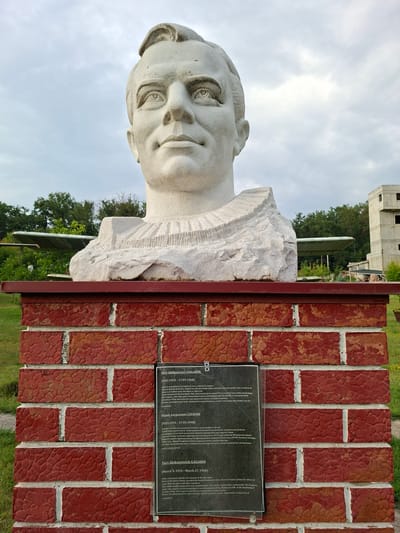 This image depicts a bust of Yuri Gagarin, the first human in space, on a brick pedestal. The bust is made of white stone and is mounted on a red brick structure. A plaque with biographical information about Gagarin is affixed to the brick pedestal, presented in Romanian, Russian, and English. Behind the bust, a partially constructed building with multiple floors and exposed concrete is visible, along with trees and a cloudy sky. The presence of what appears to be an airplane wing or similar structure in the background suggests the location might be near an airfield or aviation-related site. The general setting appears to be an outdoor park or memorial area. The scene is captured from a slightly low angle, looking up at the bust. The city of Malcoci, Moldova, is the location of this monument, specifically the House of Culture, which houses a museum dedicated to Gagarin.