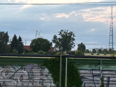 The image shows a view of a rural landscape with power lines stretching across the sky. In the middle ground, wind turbines are visible, suggesting a renewable energy source. Behind the turbines, a cluster of trees forms a dense green canopy. To the right, a tall lattice tower, likely a telecommunications tower, pierces the sky. In the foreground, a green painted fence with horizontal wooden slats is covered in graffiti. Some of the graffiti appears to be abstract shapes and lines in shades of grey and black. There is also a section of ivy growing on the fence, adding a touch of natural greenery. The sky is overcast with light clouds. The overall scene depicts a blend of industrial infrastructure and natural elements. The location is identified as Wustermark, Germany.