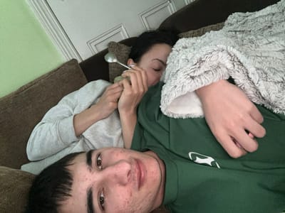 The image captures two individuals resting on a brown upholstered sofa in an interior residential setting in Dorchester, United Kingdom. In the foreground, a young male, displaying short dark hair and facial blemishes, is positioned semi-recumbent and looks directly at the camera. He wears a dark green short-sleeved top with a partial white logo. Behind him, partially obscured, a young female lies with her eyes closed, suggesting rest or sleep. She has dark hair and wears a light grey long-sleeved top. Her upper body is covered by a fluffy, light grey and white blanket. Her right hand holds a silver-colored spoon near her face. The male's left hand is visible resting on her body beneath the blanket. The background features a light green wall and a white wooden door or door frame with molding.