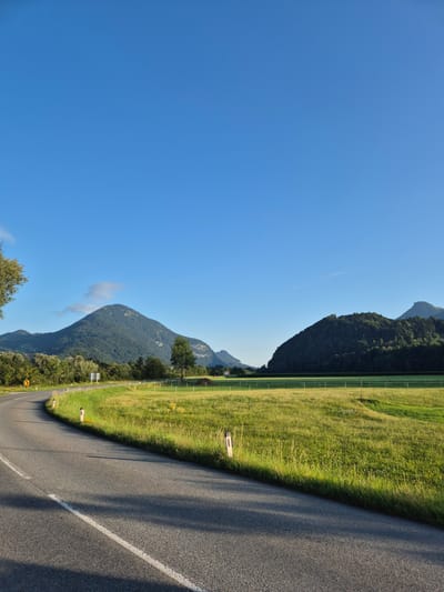 The image shows a scenic view of a curved asphalt road in Niederndorf, Austria. The road has a white line marking the edge and a broken white line in the middle. On the right side of the road, there is a vibrant green grassy field with a white fence. In the background, there are rolling hills covered with dense green trees, and in the distance, majestic mountains rise under a clear blue sky. The sunlight casts shadows on the road, indicating it is likely morning or late afternoon. The overall atmosphere is peaceful and serene, highlighting the natural beauty of the Austrian landscape.