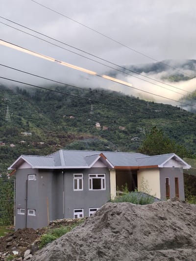 The image depicts a two-story residential building with a gray exterior and a silver corrugated metal roof, situated in Chattan Seri, India. The building features multiple white-framed windows of various sizes and a recessed entryway with wooden trim. In the foreground, a large pile of sand and construction debris is visible, suggesting ongoing development or renovation.
The background showcases a lush, green hillside dotted with smaller houses and electrical transmission towers. The upper portion of the image is dominated by a cloudy sky with streaks of light, possibly from a moving vehicle or atmospheric phenomena, suggesting a slightly overcast or misty day. The overall scene captures a residential area in a hilly region.