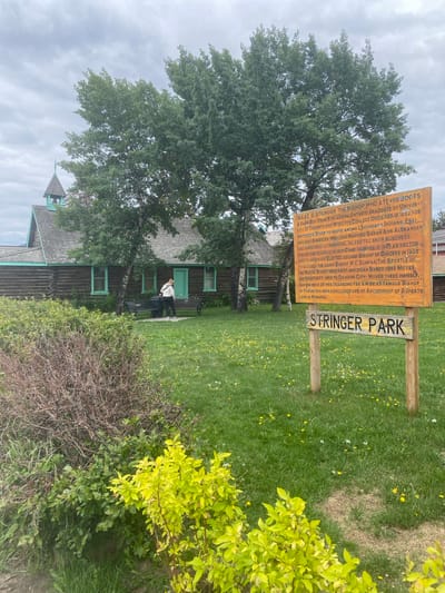In Whitehorse, Yukon, Canada, a wooden sign reading "STRINGER PARK" stands in a grassy area dotted with yellow dandelions. To the right of the sign, a larger wooden sign displays text detailing the life of Isaac O. Stringer, a bishop. Behind the signs, a rustic log building with a steeple and mint green trim is visible. Two people are standing near benches in front of the building. The sky is overcast.