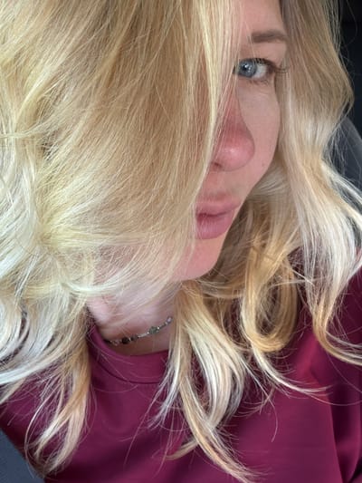 The image is a close-up, waist-up portrait of a woman with blonde hair, looking towards the right side of the frame. Her face is partially obscured by her hair, with her left eye visible. She is wearing a dark pink or magenta turtleneck shirt and a delicate beaded necklace. The background is out of focus, suggesting an indoor setting or a vehicle. There is no discernible location within Ново-Тебердинское сельское поселение, Russia.