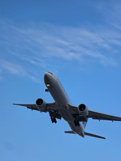 An airplane is captured from a low angle as it descends against a bright blue sky with wispy clouds. The aircraft is a large commercial jet with two engines mounted under its wings. The landing gear is extended, indicating it is in the process of landing. The front of the fuselage, nose, and cockpit windows are visible. The wings extend horizontally from the body of the plane. The scene suggests the airplane is approaching Frankfurt Airport, located near Kelsterbach, Germany.