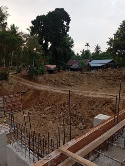 The image depicts a construction site in the city of Blangraleu, Indonesia. In the foreground, a concrete foundation with rebar reinforcement is visible, indicating ongoing construction work. Wooden formwork is in place, suggesting preparations for pouring concrete. The background reveals a landscape with cleared earth, tire tracks from heavy machinery, and piles of soil, characteristic of excavation and land leveling. An orange excavator is parked on a raised mound of earth. Further in the distance, several simple houses with tin roofs are nestled among lush green trees and palm trees, indicating a rural or semi-rural setting. The sky is overcast, suggesting it may be early morning or late afternoon. The overall scene captures a moment in the development of infrastructure within a tropical environment.