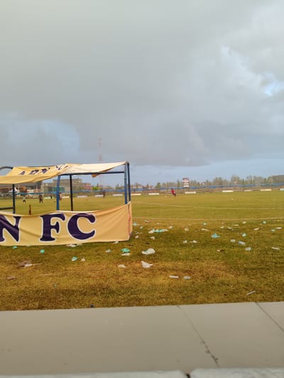 The image depicts a football field at Cibrekbaroh, Indonesia, under a cloudy sky. In the foreground, a yellow banner with purple lettering reading "N FC" is visible, partially covering a shelter structure. The field itself is green grass, strewn with scattered plastic bottles and wrappers, indicating a recent event or lack of cleanup. In the mid-ground, several individuals, appearing to be young people, are present on the field. Some are kicking a ball, while others are walking or standing. A tall communication tower is visible in the background, along with trees and buildings on the horizon. The overall scene suggests a casual football gathering or practice session on a somewhat neglected field.
