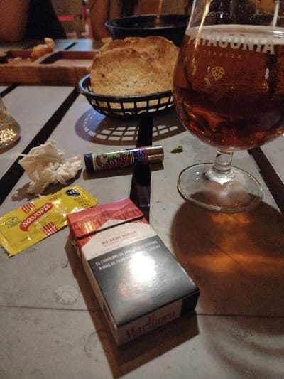 A beer in a glass sits beside a basket of bread. Next to them are a lighter with a green dragon, a crumpled napkin, a small yellow packet of Savora mustard, and a pack of Marlboro cigarettes. All are situated on a grey surface with dark shadow lines.