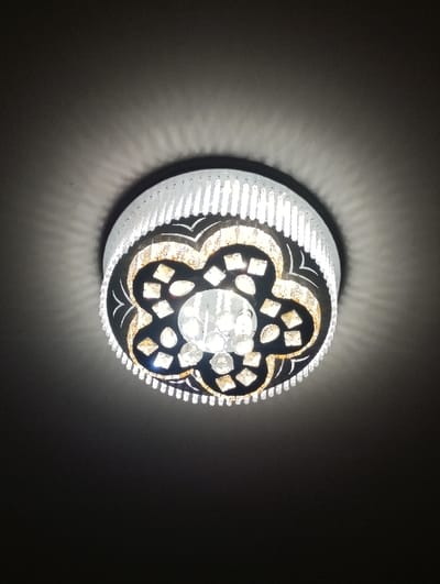 The image displays a circular, illuminated ceiling light fixture mounted against a dark, featureless ceiling in the city of Xojeli, Uzbekistan. The fixture features an outer ring composed of numerous vertical, clear, rectangular elements, likely crystal or glass. Inward from this ring, a darker, ornamental plate with a quatrefoil-like pattern contains multiple smaller, faceted, clear or amber-toned decorative crystal or glass accents. The central portion of the fixture holds several larger, spherical or teardrop-shaped clear crystal elements, serving as the primary light sources. Light emanates from the fixture, casting radial patterns onto the surrounding dark ceiling surface. No people, actions, or other objects are visible within the frame.