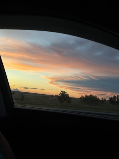 The image shows a view of a sky with clouds and a hint of sunset, seen through what appears to be a car window or sunroof. A thin strip of land with some trees is visible in the distance. The location is possibly in or near Ключи, Russia.