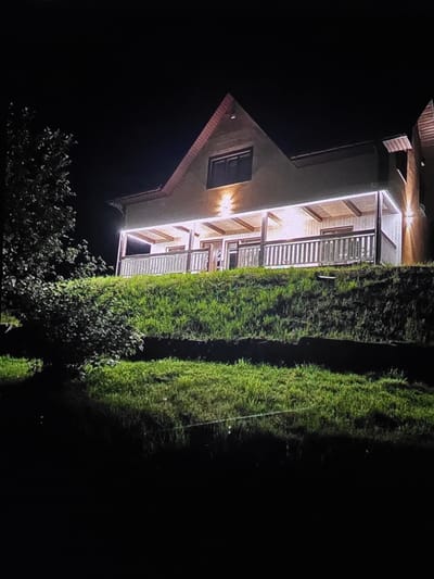 The image depicts a nighttime scene featuring a multi-story house with illuminated windows and a porch. The house has a light-colored facade and a red-tiled roof. Adjacent to the house are two tall, lush green hedges or trees, creating a strong vertical element in the composition. In the foreground, there are visible trees and foliage, which are darker and less distinct due to the low light conditions. The overall setting appears to be a residential area, possibly a garden or backyard, in Dubno, Ukraine. The time of day is clearly night, indicated by the darkness of the sky and the artificial lighting from the house. There are no people visible in the image, and the focus is on the architecture and the natural elements surrounding it. The lighting highlights the details of the house's porch, with visible wooden beams and railings. The tall hedges are well-lit by an unseen light source, creating a contrast with the dark surroundings. There is no visible text in the image. The scene conveys a sense of quietude and domesticity, with the illuminated house standing out against the night.