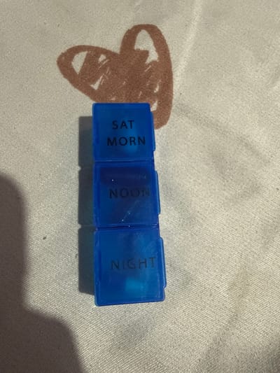 A stack of three blue plastic pill organizers is placed on a beige fabric surface with a brown heart-shaped stain. Each organizer is labeled with text indicating a time of day. The top organizer reads "SAT MORN", the middle organizer reads "NOON", and the bottom organizer reads "NIGHT". The image does not provide any information about the city of Manila, Philippines, or any specific details regarding people, actions, or interactions.