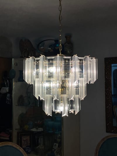 A chandelier hangs in a room in Clearwater, United States of America. The chandelier has clear glass panes in a rectangular shape. The chandelier is attached to a gold colored chain. The room contains decorative items and mirrors.
