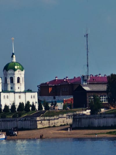 The image depicts a waterfront scene in Yeniseysk, Russia, featuring a prominent white building with a green dome, likely a church or monastery, on the left side. Adjacent to it is a complex of buildings with red roofs. A telecommunications tower stands tall in the background. The foreground shows a river with a small white and blue boat docked on the shore. A concrete embankment separates the buildings from the sandy riverbank, where several individuals are visible, some standing and others walking. Trees are scattered throughout the scene, and the sky is a clear blue. The overall atmosphere suggests a sunny day in a historic Russian city.