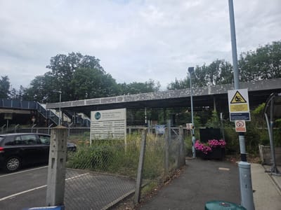 The image shows a street scene in Elmbridge, United Kingdom. In the foreground, a black car is parked next to a chain-link fence. A concrete post stands in front of the fence. Behind the fence, there is a sign for "Hinchley Wood Practice" which offers various medical services including orthodontics, physiotherapy, chiropody/podiatry, psychotherapy, Pilates, and holistic therapy. There is also a sign with a warning symbol of a person falling, stating "In the interest of your safety please take extra care in wet weather and icy conditions." Further down the street, there is a "No Smoking" sign. In the background, there is a bridge and trees. The sky is overcast.