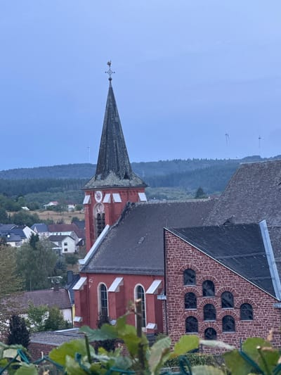 The image depicts the parish church of St. Martin in Wadern, Germany. The church features a tall, dark steeple topped with a weather vane shaped like a rooster. The main body of the church is painted red and has arched windows. Adjacent to the steeple is a section of the church constructed from red brick, with a series of smaller arched windows. In the background, a village with houses and trees is visible, situated on a hillside. Further in the distance, a dense forest covers rolling hills, and several wind turbines can be seen on the horizon. The sky is overcast with a pale blue hue. There are no people or animals visible in the image.