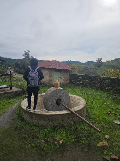 The image depicts an outdoor scene in a rural or semi-rural area of Moqrisset, Morocco, under an overcast sky. In the foreground, a person is standing with their back to the viewer, wearing a dark hooded jacket, dark pants, and athletic shoes, and carrying a grey backpack. They are positioned on muddy green ground, facing a traditional grinding stone mechanism. This mechanism consists of a large, rough-hewn circular stone millstone set vertically within a circular concrete basin. A dark metal rod protrudes from the center of the millstone, extending diagonally downwards to the right within the basin. A small, orange-capped plastic bottle rests upright on top of the millstone.

Behind the grinding stone, a small, single-story building with stone walls and a reddish-brown corrugated metal roof is visible. It features a light green wooden door in its facade. A low stone wall extends from the right side of this building. To the left of the building, a dirt path winds into the distance, where indistinct figures of other individuals can be seen. The background features undulating green hills covered with trees and sparse vegetation, characteristic of a natural landscape. The ground in the foreground is wet and muddy, with patches of vivid green grass.