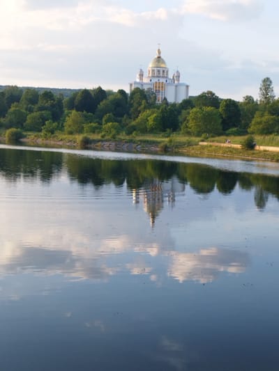 The image shows the Basilica of the Assumption of the Blessed Virgin Mary in Novyi Rozdil, Ukraine, with its reflection in the calm waters of a river in the foreground. The basilica, a large white building with a golden dome, is situated on a hill amidst lush green trees. The sky above is partly cloudy, with soft sunlight illuminating the scene. A few people are visible walking along a path by the riverbank, adding a touch of human presence to the otherwise tranquil landscape. The reflection of the basilica and the surrounding trees in the water creates a symmetrical and visually appealing composition. The overall atmosphere is serene and picturesque, showcasing the beauty of the architectural landmark and its natural surroundings.