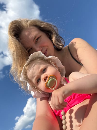 In this close-up, low-angle shot, a woman and a baby are positioned against a backdrop of a bright blue sky with scattered white clouds. The woman, with blonde hair, is looking down at the baby. The baby, wearing a white bonnet and a pink swimsuit, has a pacifier in its mouth and a string of wooden beads draped over its hand. The woman is wearing a dark green swimsuit top. The perspective suggests they are outdoors, possibly at a beach or park, given the bright sunlight and open sky. The location is not identifiable within the city of Колокша, Russia from the visual information provided.