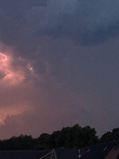 In the city of Putten, Netherlands, at dusk, a storm approaches. Dark, heavy clouds dominate the sky, with streaks of lightning illuminating the roiling masses in shades of pink and white. Below the stormy sky, a dense line of dark green trees stretches horizontally across the frame. In the foreground, the dark, tiled roofs of houses are visible, with one roof featuring two dormer windows. The scene captures the dramatic transition from day to night under the threat of a thunderstorm.
