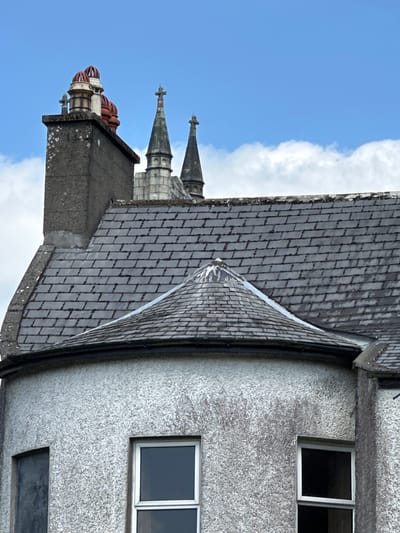This is a vertical, eye-level outdoor shot of a building with a slate roof and a textured grey facade. The building appears to be in Baltinglass, Ireland. The roof is made of dark grey slate tiles arranged in a diamond pattern, with a prominent, steeply pitched section in the foreground that meets another section at an angle. A metal flashing runs along the edge of the angled roof.
The building's facade is a rough, speckled grey, with two white-framed windows visible. The upper window is rectangular and has two panes; the lower window is also rectangular but appears to be a single pane. The lighting suggests it is daytime, likely late morning or afternoon, given the brightness and the shadow cast by the roof. The sky in the background is a vibrant blue with scattered white clouds.
In the background, to the left of the main building, are architectural elements that suggest a more ornate, possibly older structure attached or nearby. These include spires with pointed tops and ornate decorative elements, some appearing to be red and white. There is no text visible in the image. The scene evokes a sense of historical architecture and a pleasant, clear day.