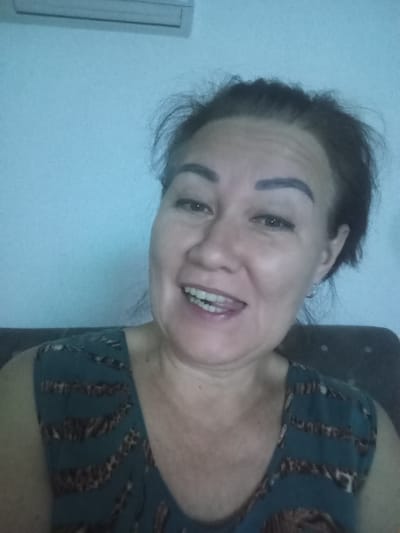 A woman is shown from the chest up in a selfie-style image. She has dark hair pulled back and appears to be wearing makeup. Her mouth is open in a wide smile, showing her teeth. She is wearing a dark teal top with a leopard print pattern. The background is a plain light blue wall with what appears to be an air conditioning unit above her. The image is not of Yukariyuz, Uzbekistan.