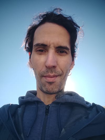 A low-angle portrait of a man with dark hair and a stubbled beard, wearing a blue hooded sweatshirt, against a clear, light-blue sky. The light source appears to be behind and slightly below the man, creating a bright halo effect around his hair. The man is looking directly at the camera with a neutral expression. The picture location is not discernible from the image. Therefore, the location cannot be determined.