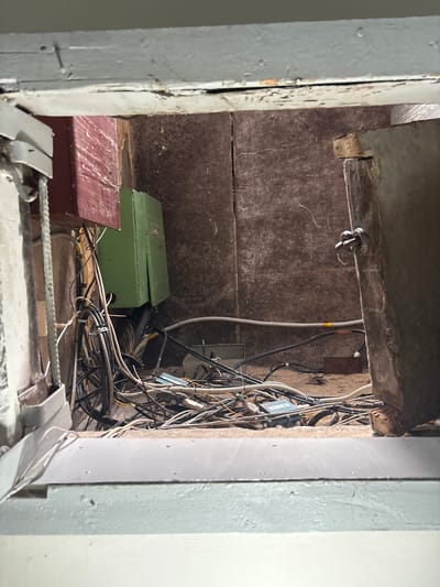 The image captures a cluttered interior space viewed through an opening. The scene is dominated by a dense tangle of electrical wires and cables, suggesting a utility area or a place with extensive networking equipment. A green rectangular box, possibly an electrical panel or junction box, is prominently positioned amongst the wires. To the left, a portion of a red-painted surface is visible, and to the right, a rustic metal door with a latch mechanism is ajar. The walls appear to be rough and textured, with visible cracks. The overall impression is one of disarray and neglect, with exposed wiring and unfinished elements. The context provided indicates this scene is located in Tutaev, Russia.