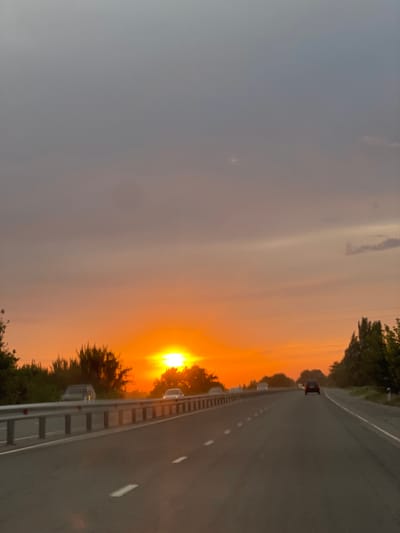 The image depicts a view from a vehicle traveling on a paved road towards a vibrant sunset. The sky is dominated by hues of orange and gray, with the sun, a bright yellow orb, partially obscured by trees on the horizon. The road has white lane markings and is bordered by a metal guardrail on the left. Several vehicles are visible on the road, including cars in the distance and one in the foreground. The environment appears to be rural, with trees and vegetation lining the sides of the road. The scene suggests a travel scene during dusk in Keŋ-Buluŋ, Kyrgyzstan.